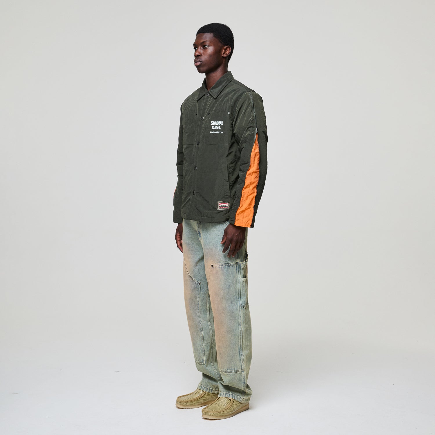 Person wearing a green Criminal Damage jacket with orange sleeves and light blue jeans on a white background