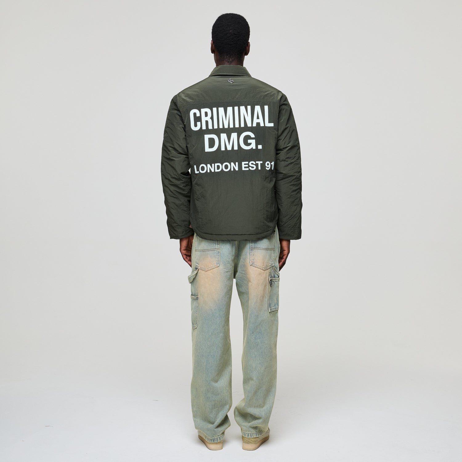 Person wearing a green jacket with 'Criminal Damage' text on the back, standing against a white background