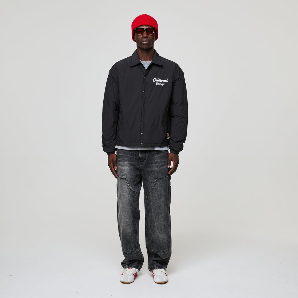 Person wearing a black Criminal Damage jacket with text, gray jeans, and a red cap on a white background