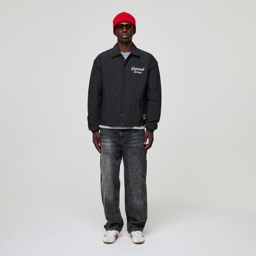 Person wearing a black Criminal Damage jacket with text, gray jeans, and a red cap on a white background