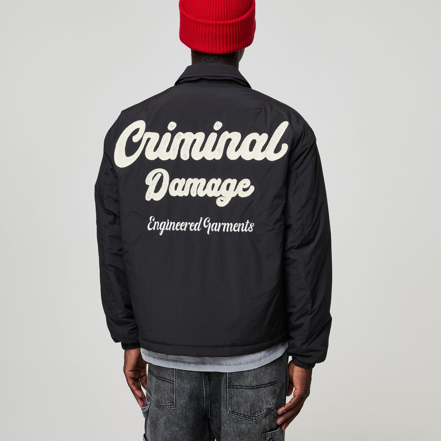Person wearing a black Criminal Damage  jacket with 'Criminal Damage' text on the back, standing against a plain background.