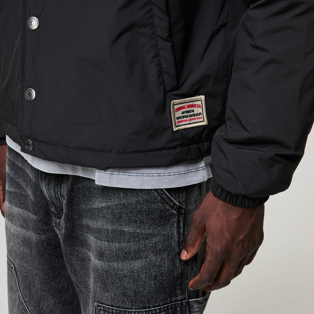 Person wearing a black Criminal Damage jacket with a brand patch on a neutral background