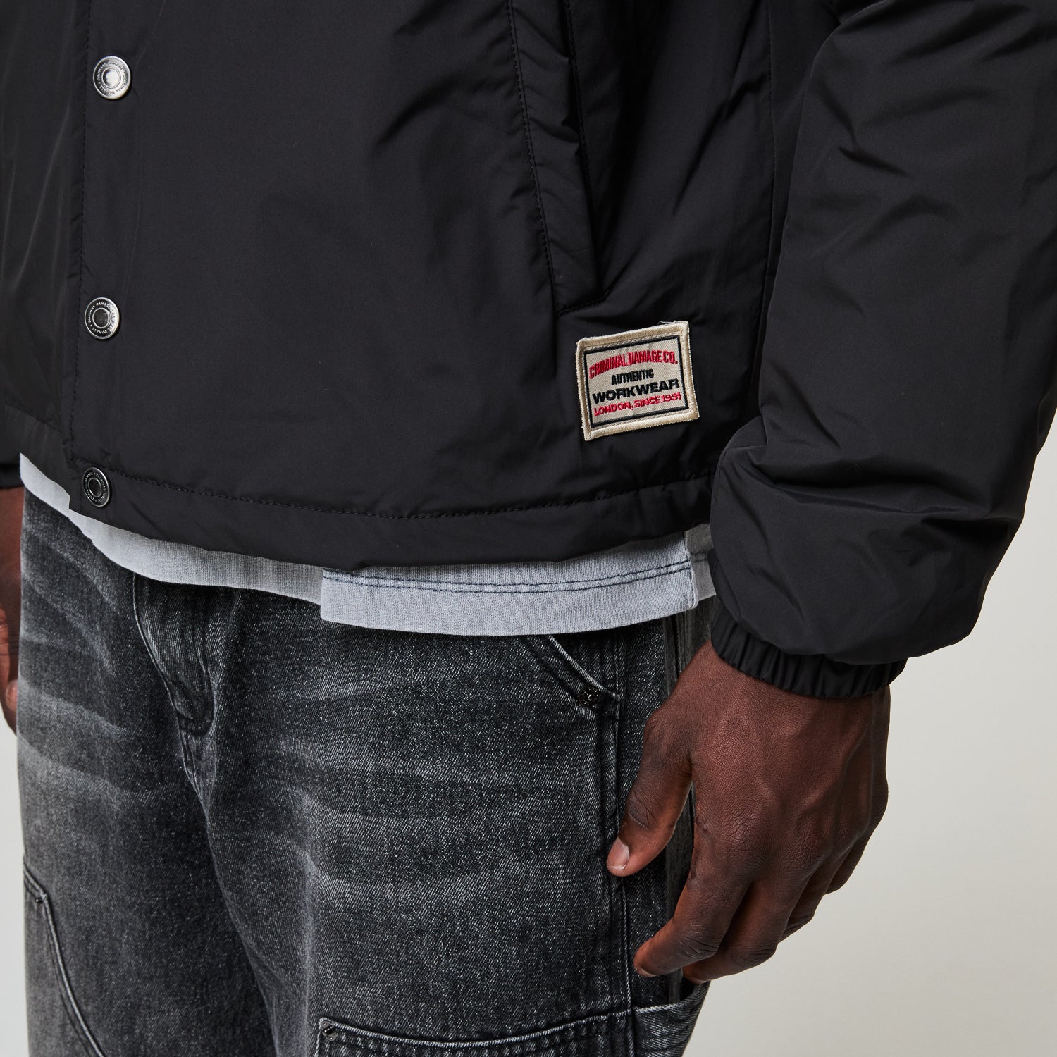 Person wearing a black Criminal Damage jacket with a brand patch on a neutral background