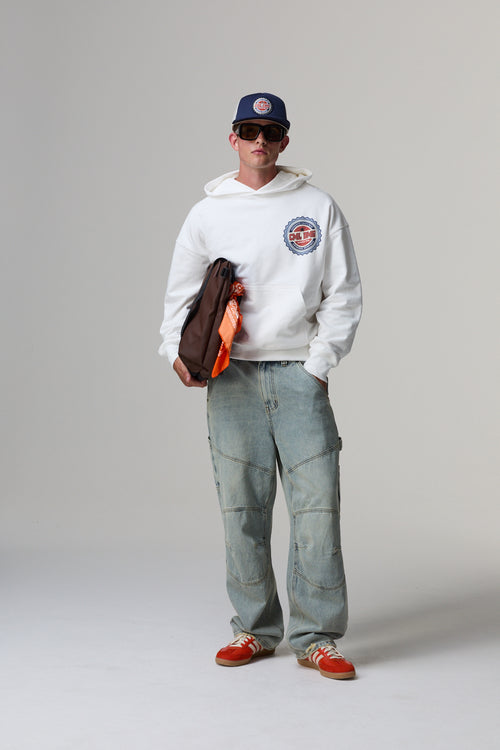 Model wearing a white Criminal Damage hoodie with a logo, light blue jeans, and red shoes on a grey background