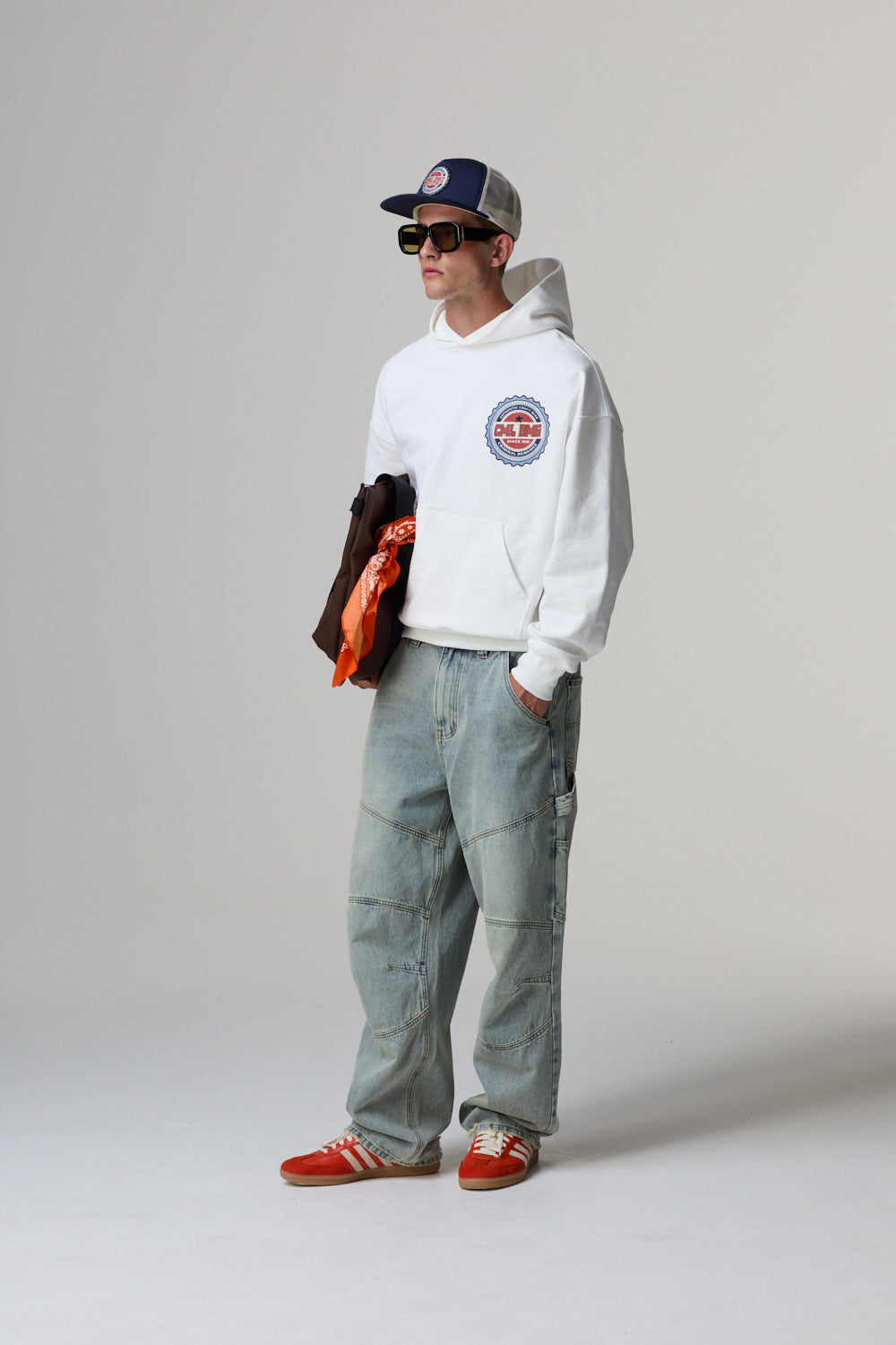 Model wearing a white Criminal Damage hoodie, grey pants, and red shoes on a grey background