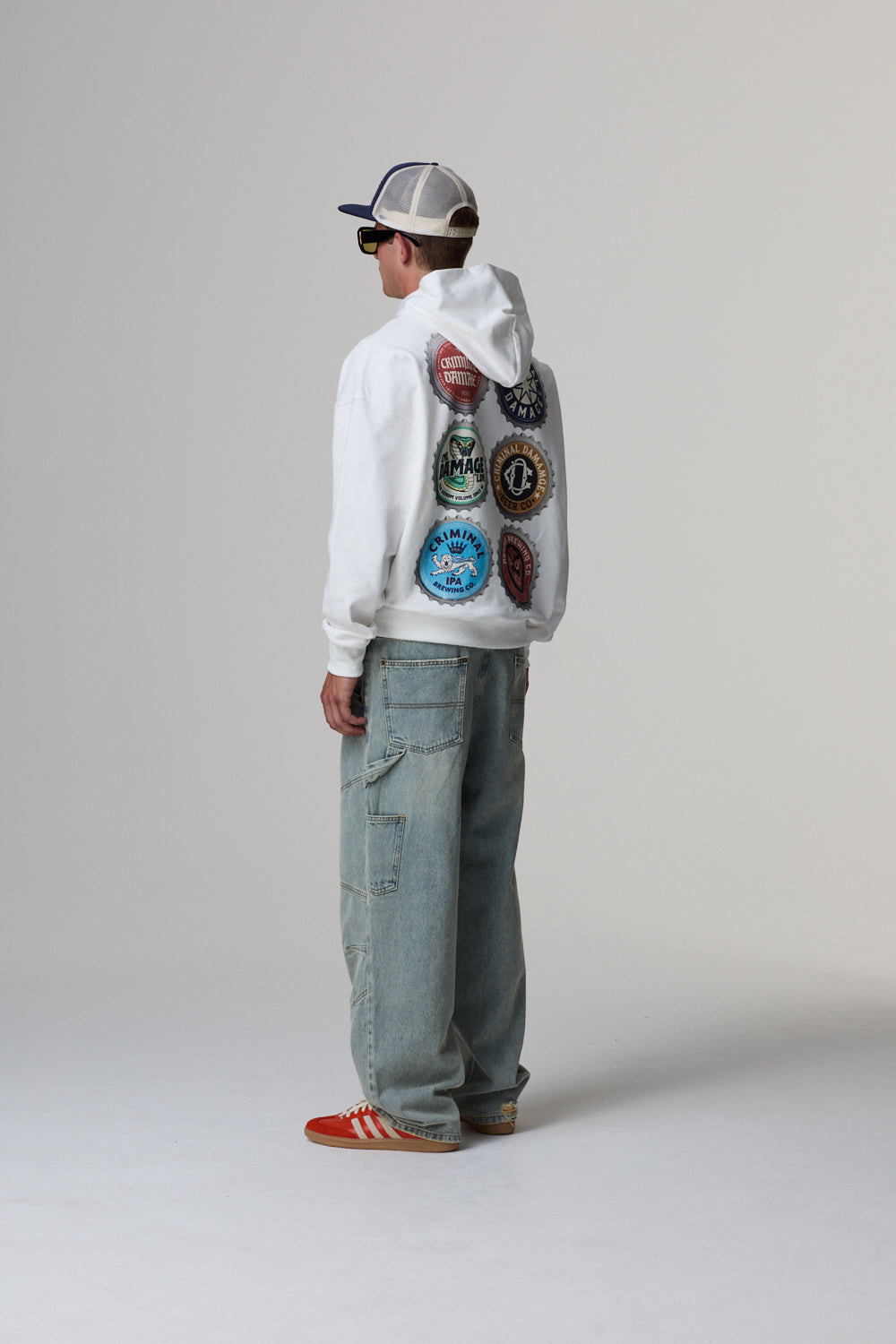 Model wearing a white Criminal Damage hoodie with colourful graphic on the back, standing against a grey background.