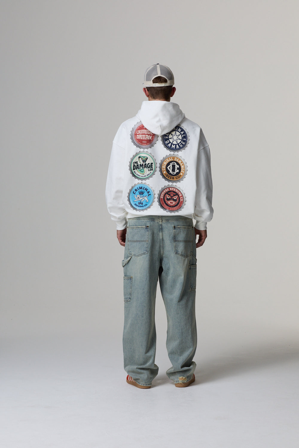 Model wearing a white Criminal Damage hoodie with colourful graphic and light blue jeans on a grey background