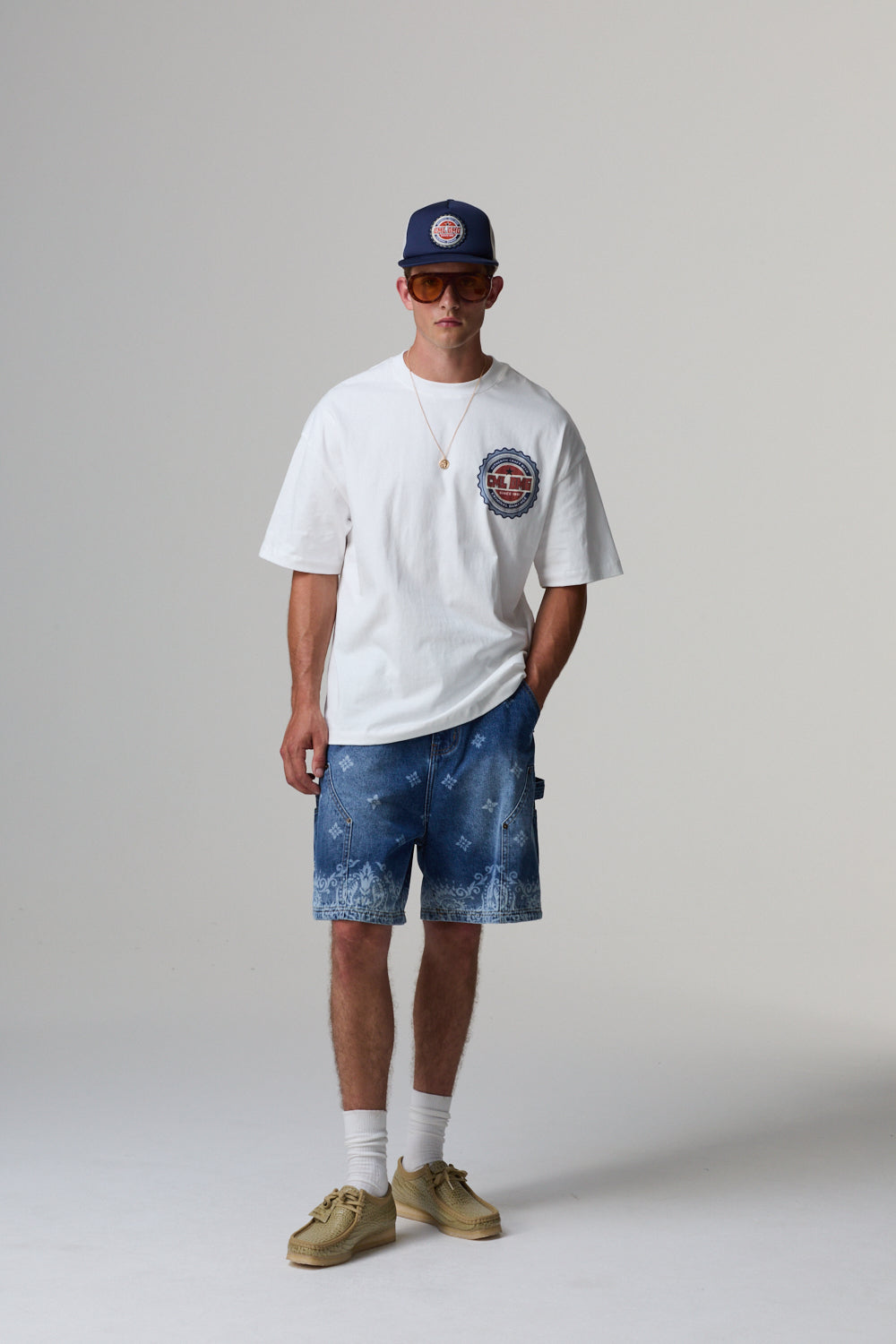 Model wearing a white t-shirt with a logo, blue shorts, and a cap on a plain background