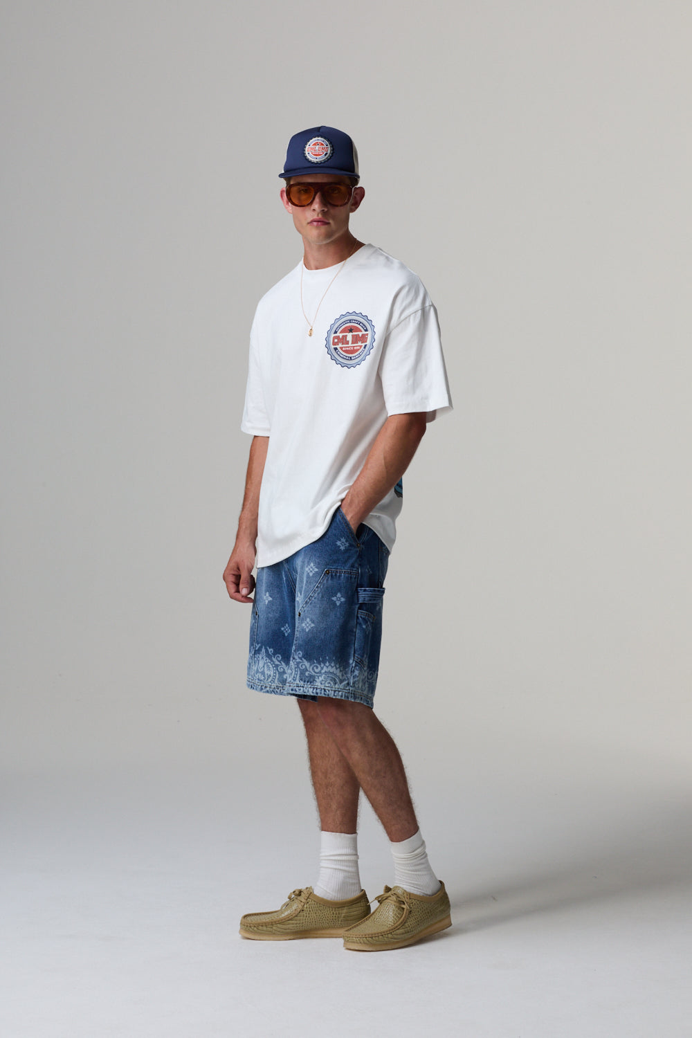 Model wearing a white t-shirt, blue shorts, beige shoes, and a navy cap on a plain background