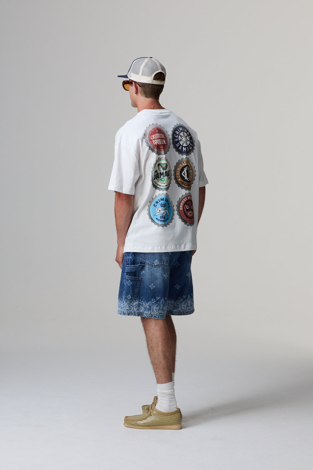 Model wearing a white t-shirt with colourful designs on the back, blue shorts, and beige shoes against a plain background.