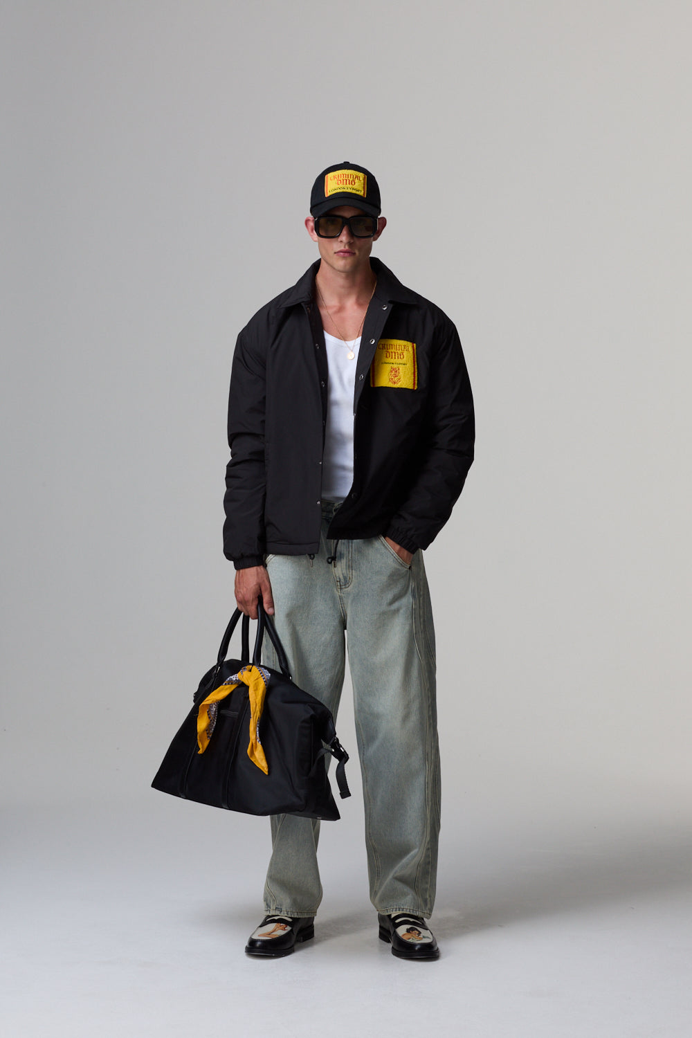 Model wearing a black Criminal Damage jacket with yellow accents, holding a black bag with yellow details, on a grey background.