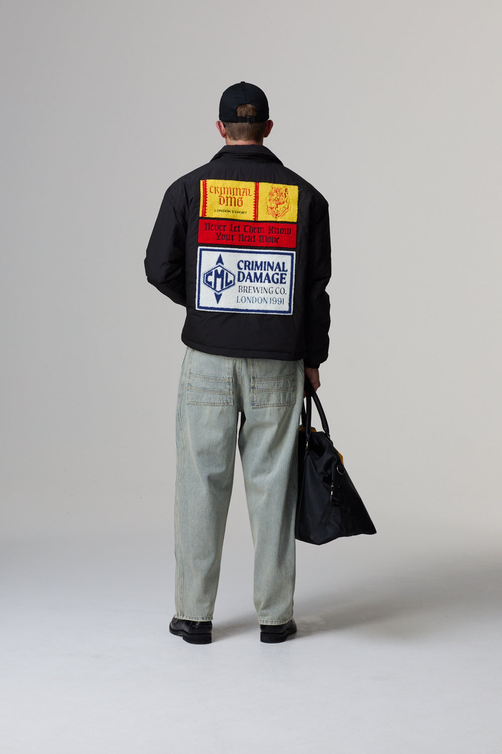 Model wearing a Criminal Damage black jacket with colourful labels on the back, holding a black bag.