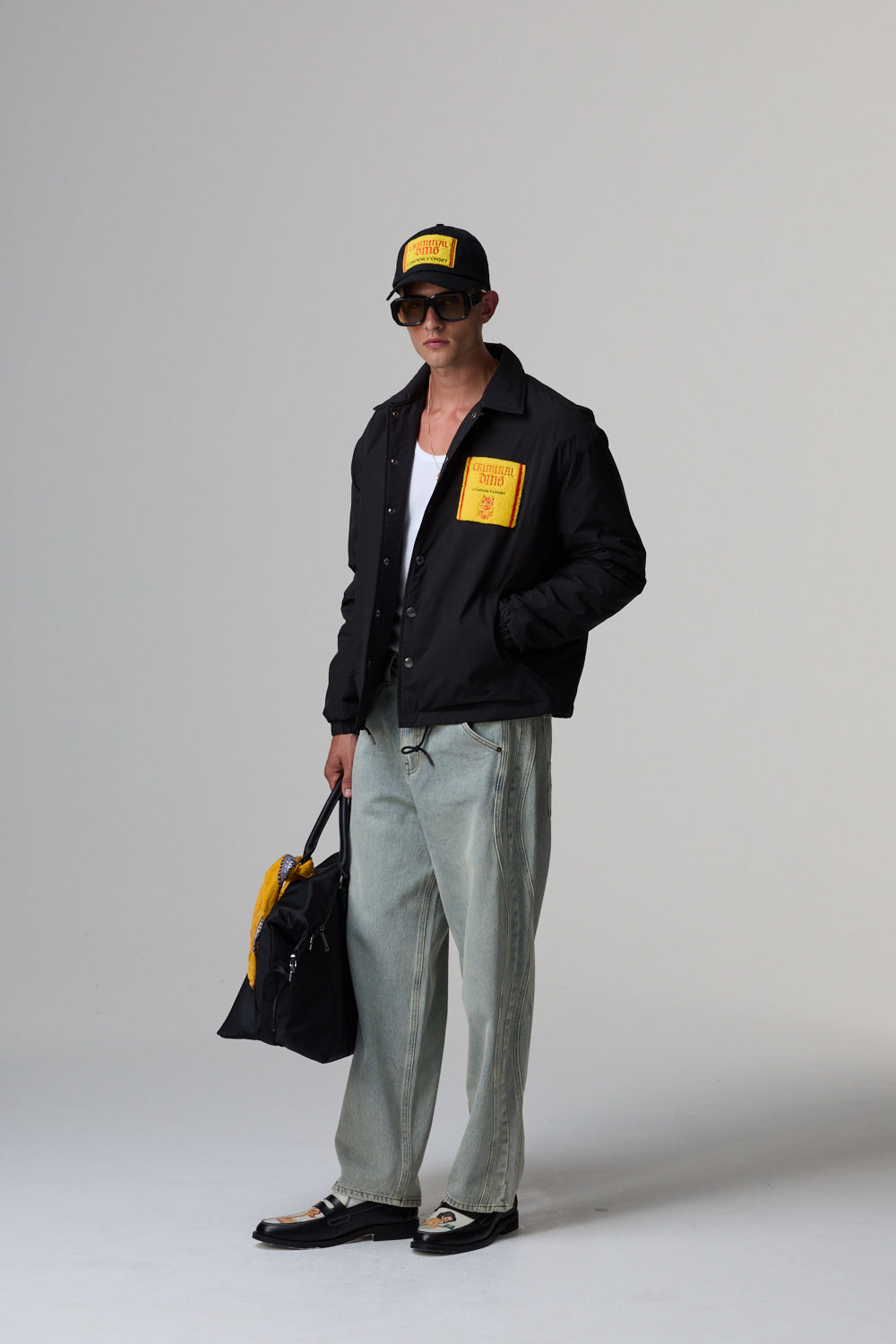 Model wearing a black Criminal Damage jacket with a yellow patch, light grey pants, and a black cap on a grey background