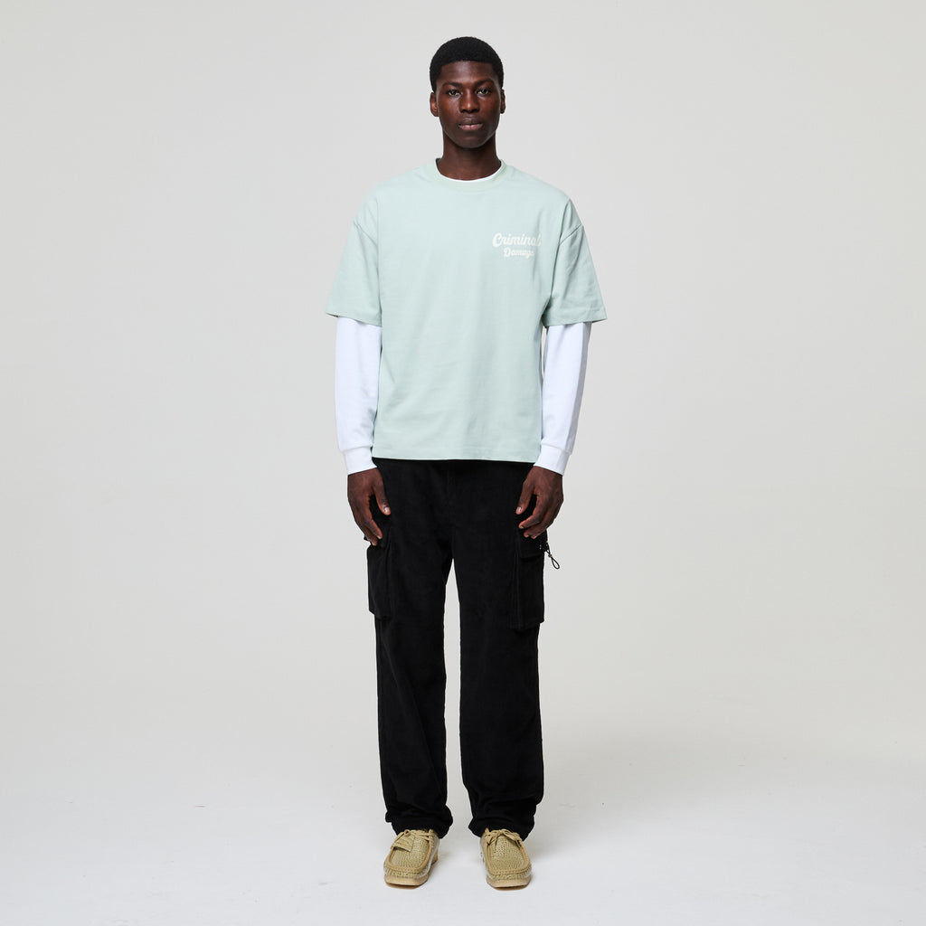 Person wearing a light green t-shirt with a white logo, black pants, and beige shoes on a plain background