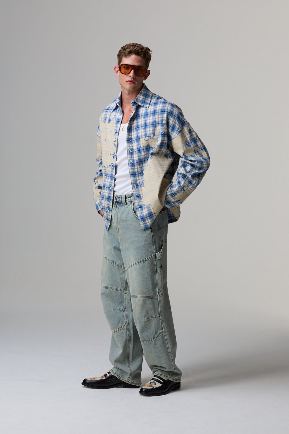 Man wearing a blue plaid Criminal Damage shirt over a white shirt, light blue pants, and black shoes on a grey background