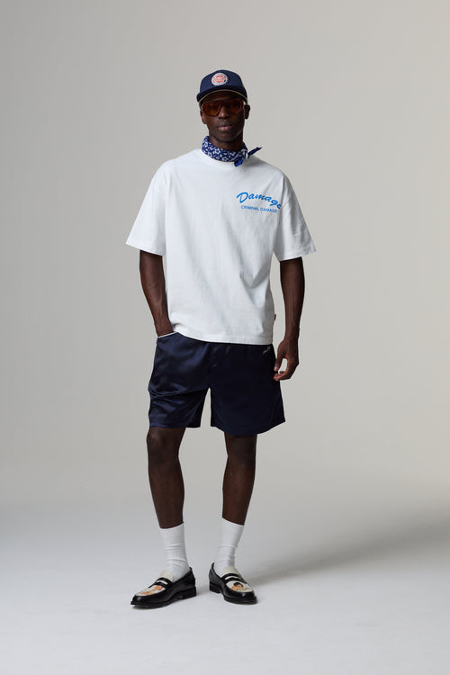 Model wearing a white Criminal Damage t-shirt with blue text, navy shorts, and a cap on a grey background