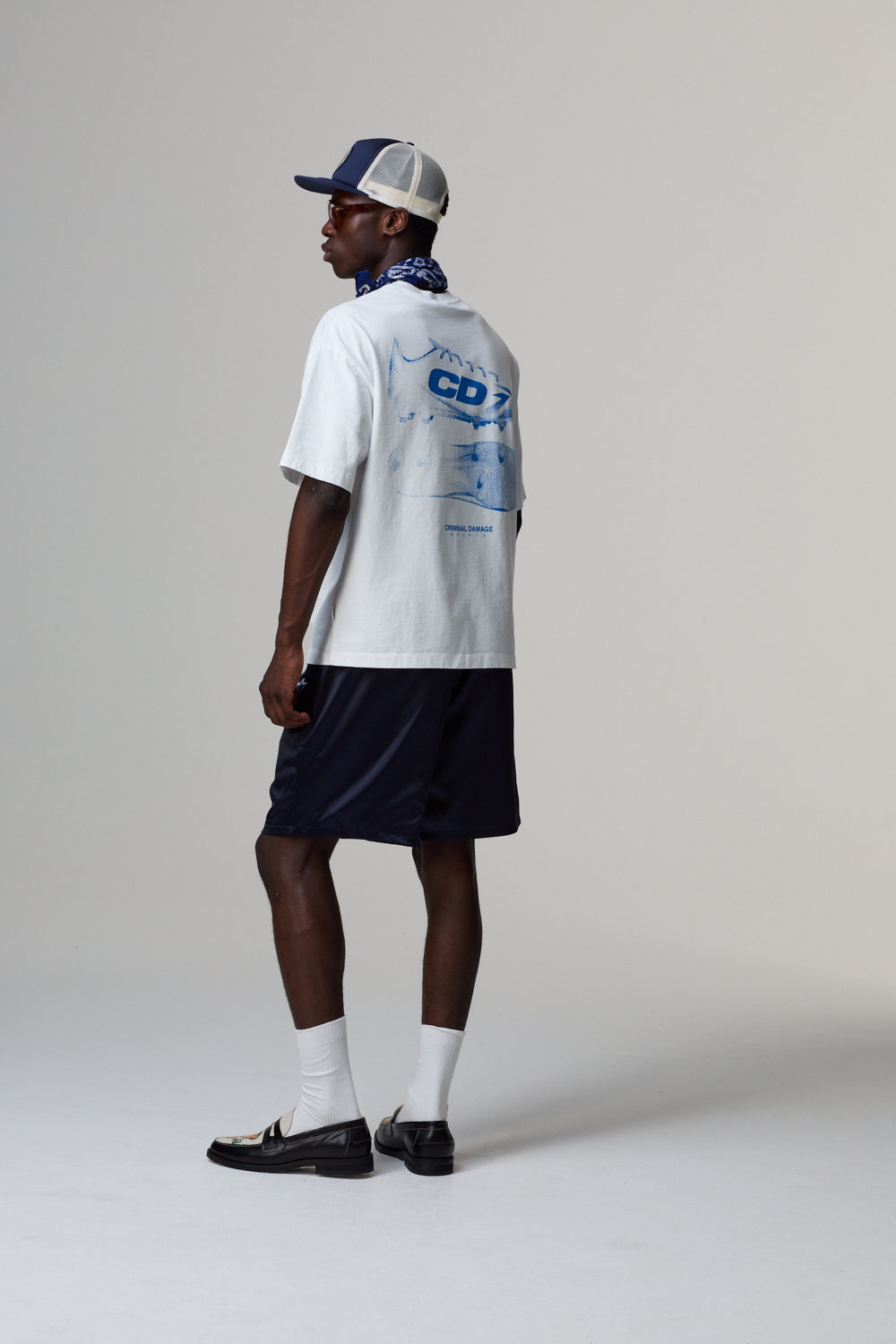 Model wearing a white Criminal Damage t-shirt with blue graphics, navy shorts, and a cap on a grey background