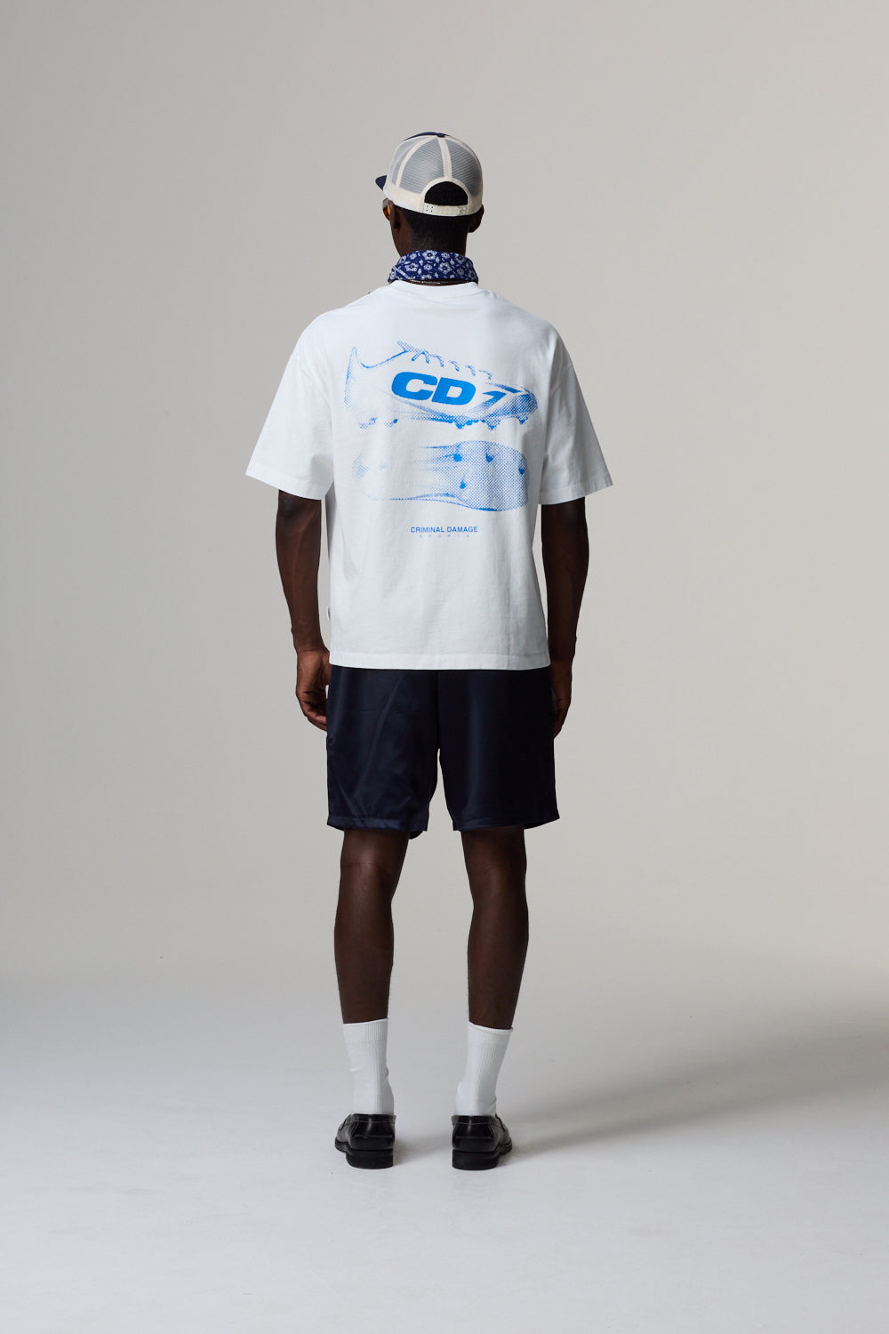 Model wearing a white Criminal Damage t-shirt with blue text and graphics, black shorts, and a cap on a grey background