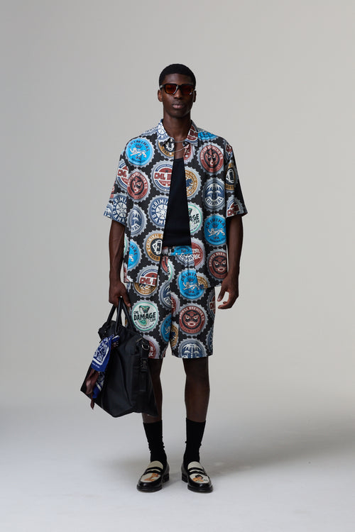 Man wearing a Criminal Damage colourful patterned outfit with sunglasses and a bag on a grey background