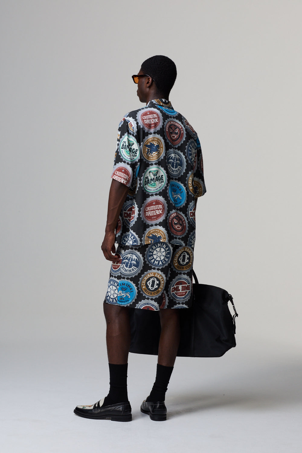 Model wearing a patterned Criminal Damage shirt and shorts, holding a black bag on a grey background