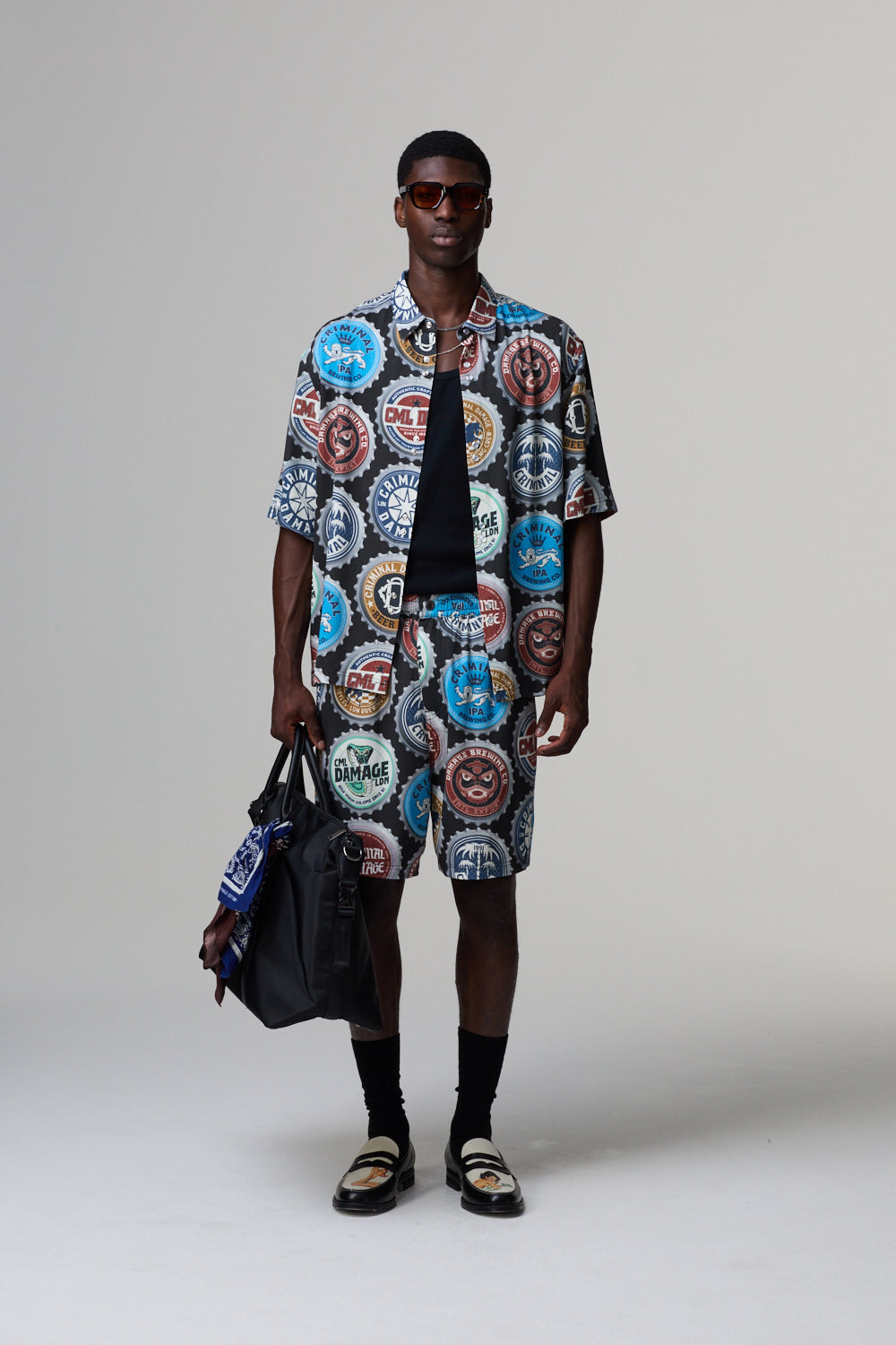 Model wearing a colourful Criminal Damage patterned shirt and shorts with a black bag on a grey background
