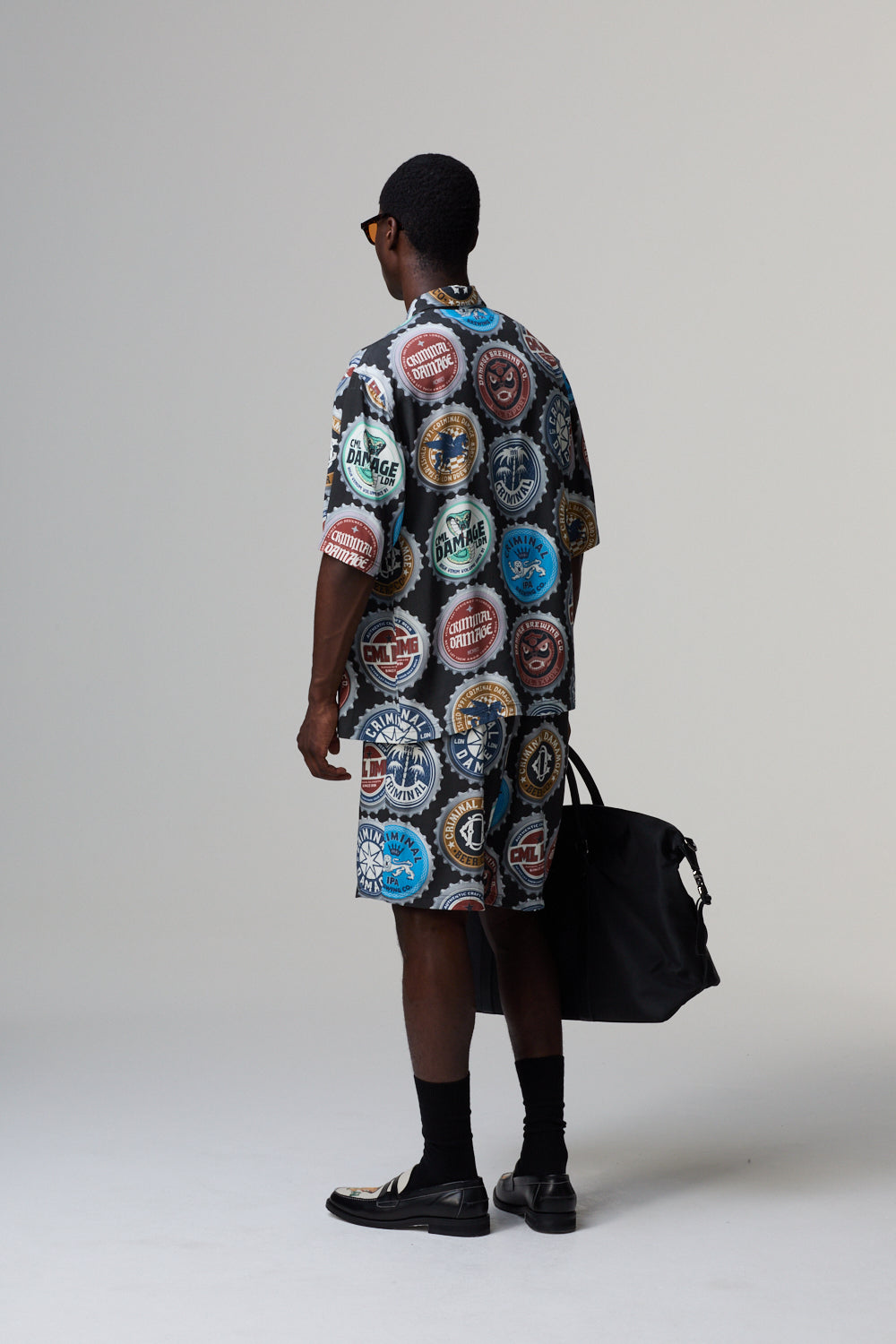 Model wearing a Criminal Damage patterned shirt and shorts, holding a black bag on a grey background
