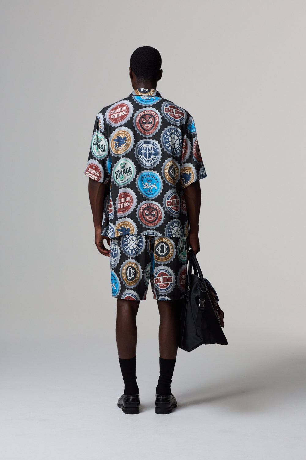 Model wearing a colorful Criminal Damage graphic outfit holding a black bag on a grey background