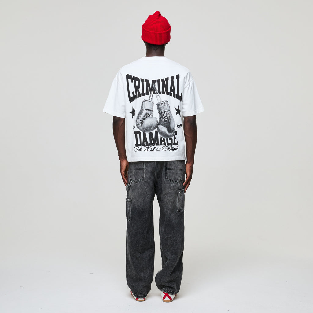 Person wearing a white t-shirt with 'Criminal Damage' text and graphics, black pants, and red beanie on a plain background
