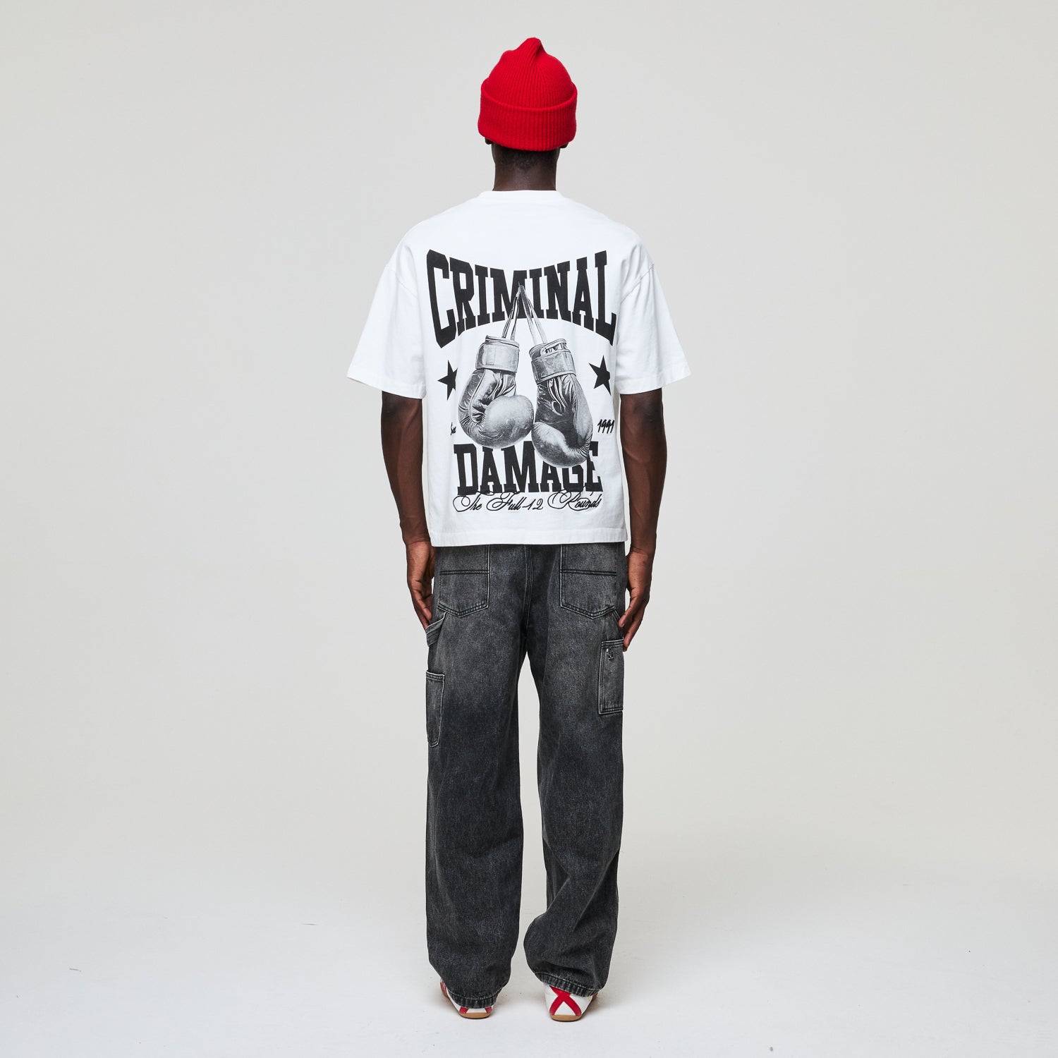 Person wearing a white t-shirt with 'Criminal Damage' text and graphics, black pants, and red beanie on a plain background