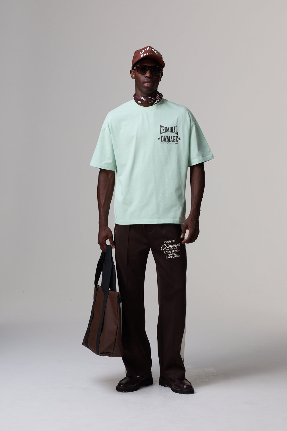 Model wearing a Criminal Damage light green t-shirt and brown pants with a cap, holding a bag on a plain background