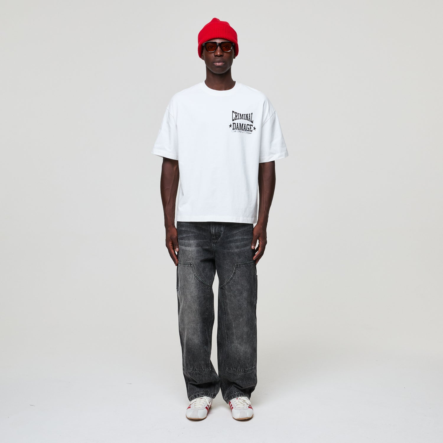 Person wearing a white Criminal Damage t-shirt, gray jeans, and a red beanie on a plain background