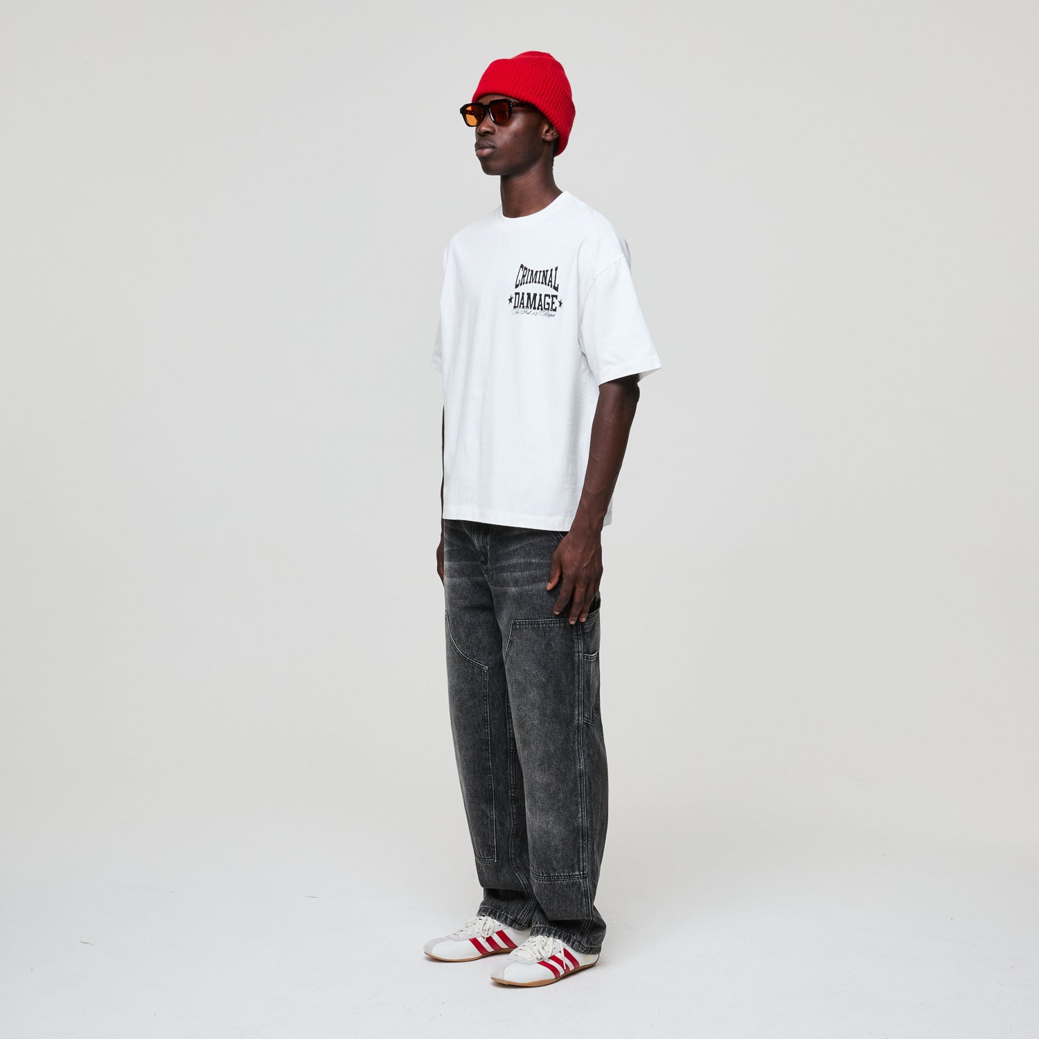 Person wearing a white Criminal Damage t-shirt, red beanie, and gray jeans on a white background