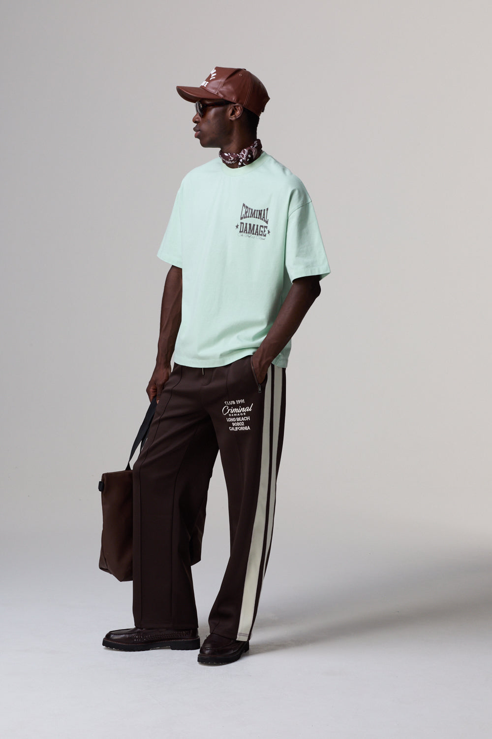Model wearing a Criminal Damage light green t-shirt and brown pants with white stripes on a plain background