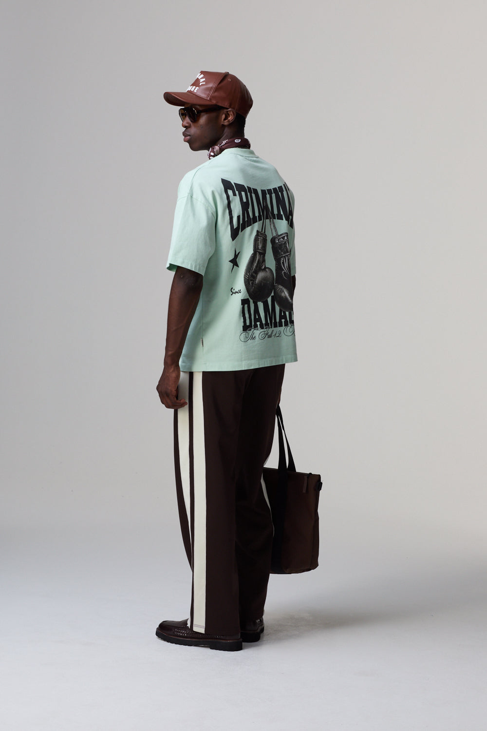 Model wearing a Criminal Damage light green t-shirt with graphic design, dark pants, and brown cap on a plain background