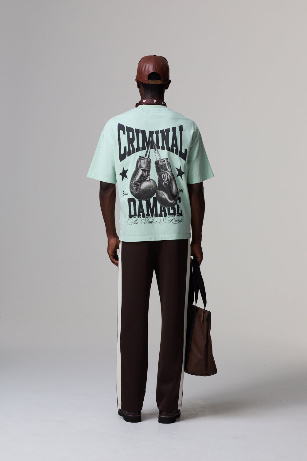 Model wearing aCriminal Damage light green t-shirt with graphic design and text, standing against a plain background.