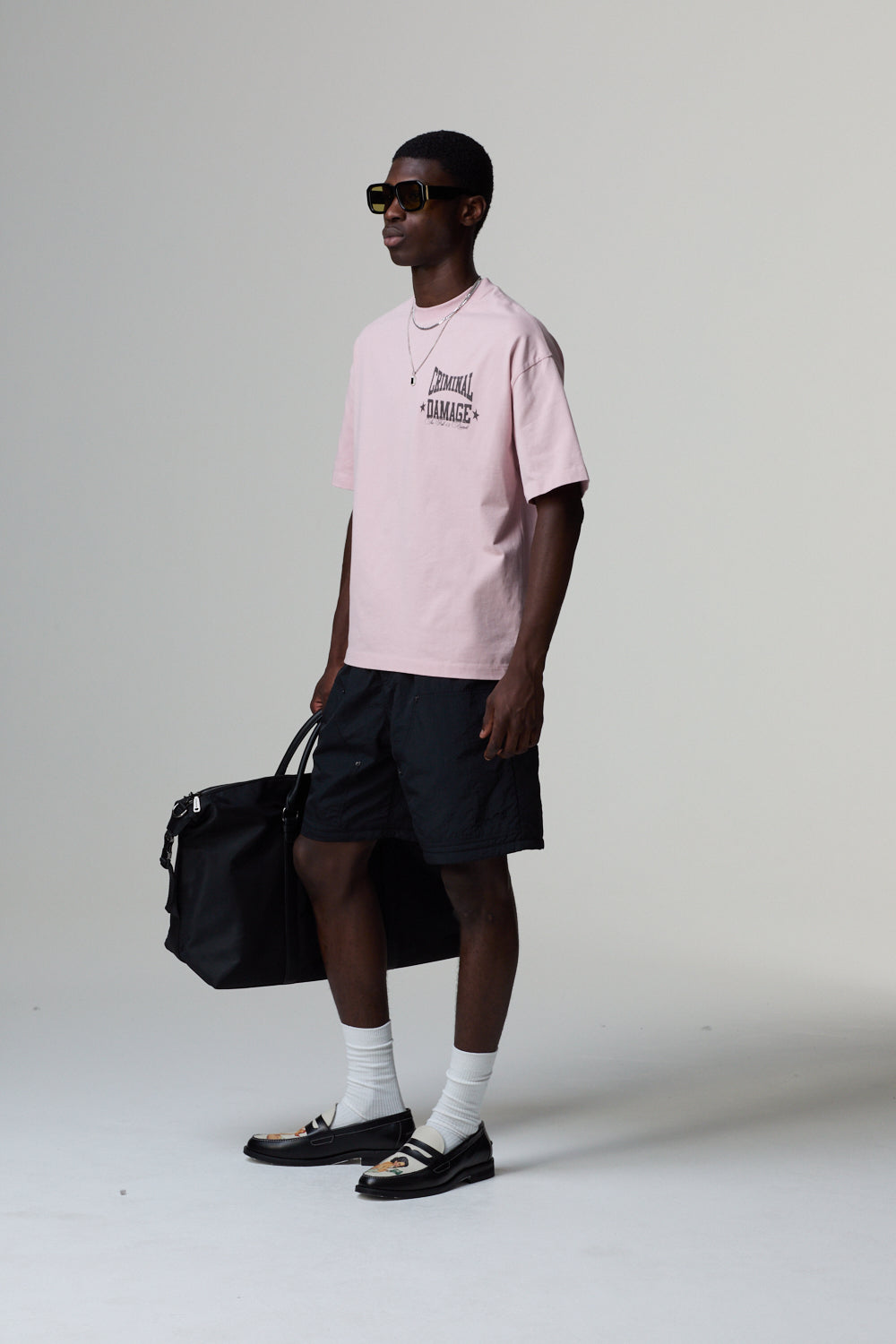 Model wearing a Criminal Damage lilac t-shirt and black shorts, holding a black bag on a white background
