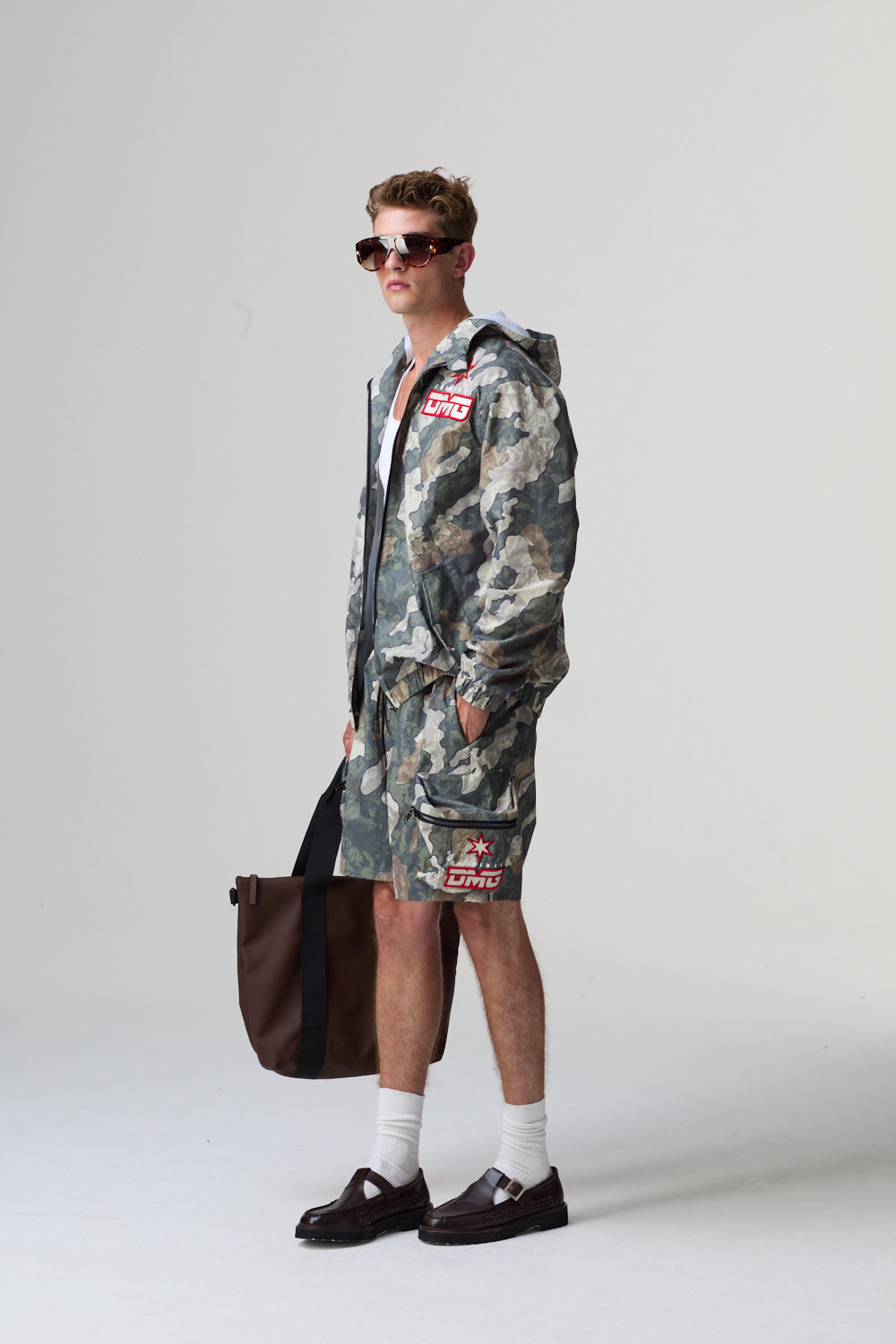 Model wearing a camouflage Criminal Damage outfit with sunglasses and a bag on a grey background