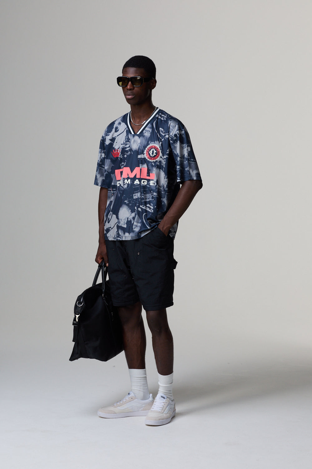 Model wearing a patterned Criminal Damage jersey and shorts holding a bag on a grey background