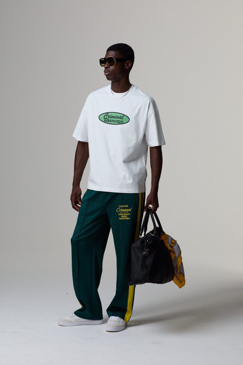 Model wearing a white a Criminal Damage t-shirt, green pants, and holding a black bag on a grey background