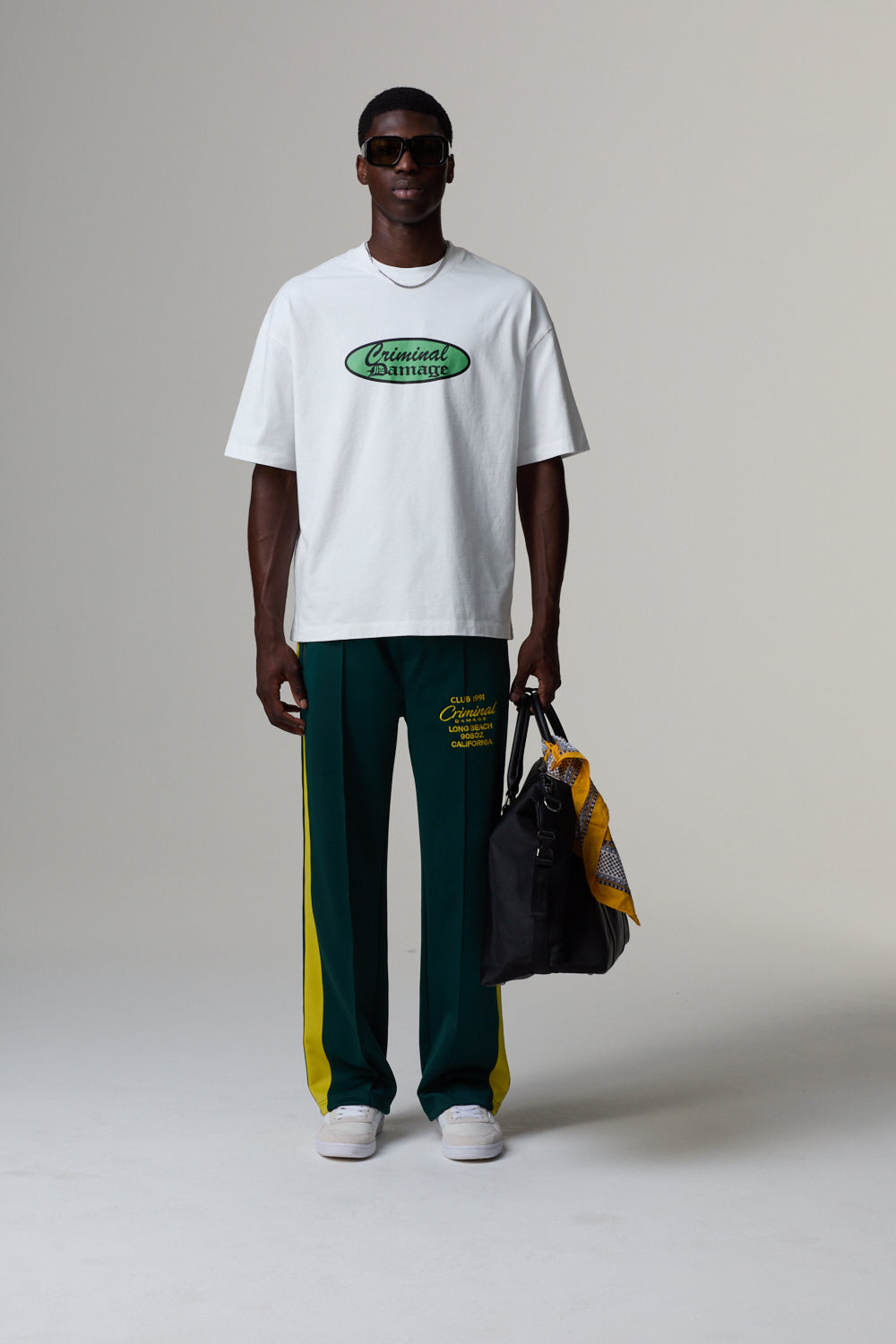 Model wearing a white Criminal Damage t-shirt with a logo and green pants, holding a black bag on a grey background