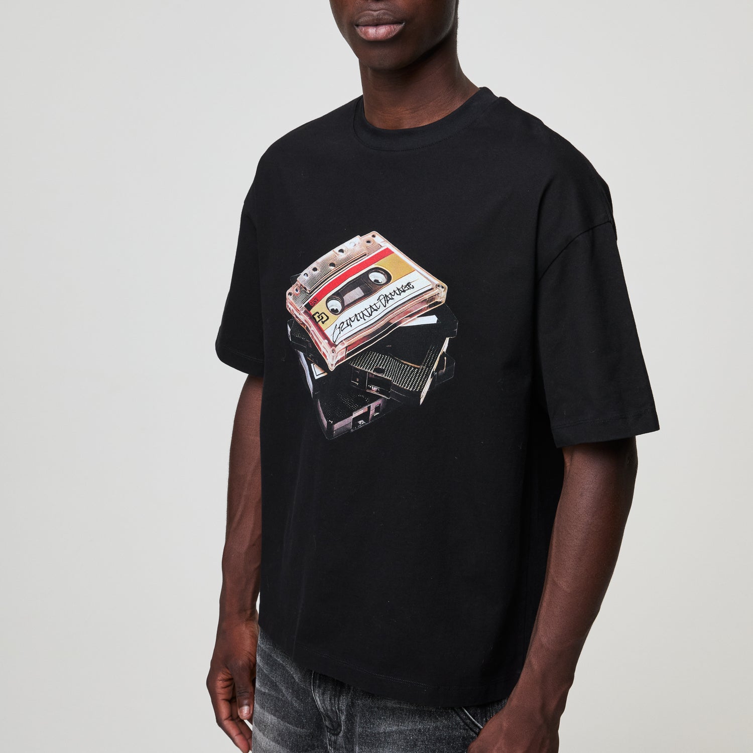 Person wearing a black Criminal Damage  t-shirt with a cassette tape graphic on a plain background