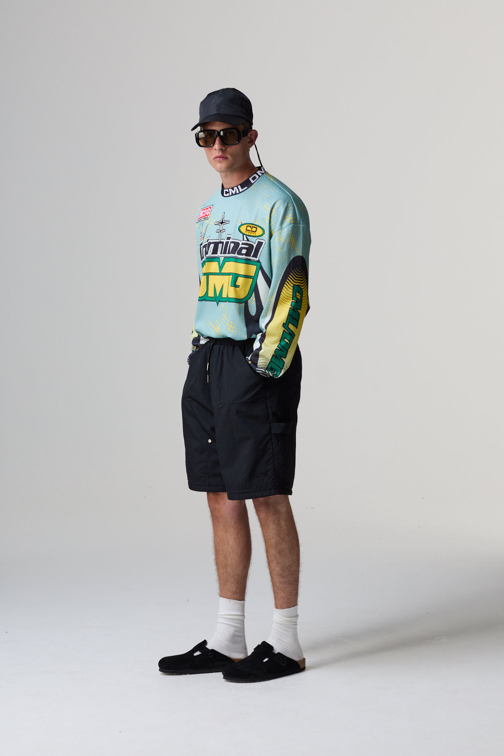 Model wearing a colourful Criminal Damage jersey, black shorts, and a cap on a grey background