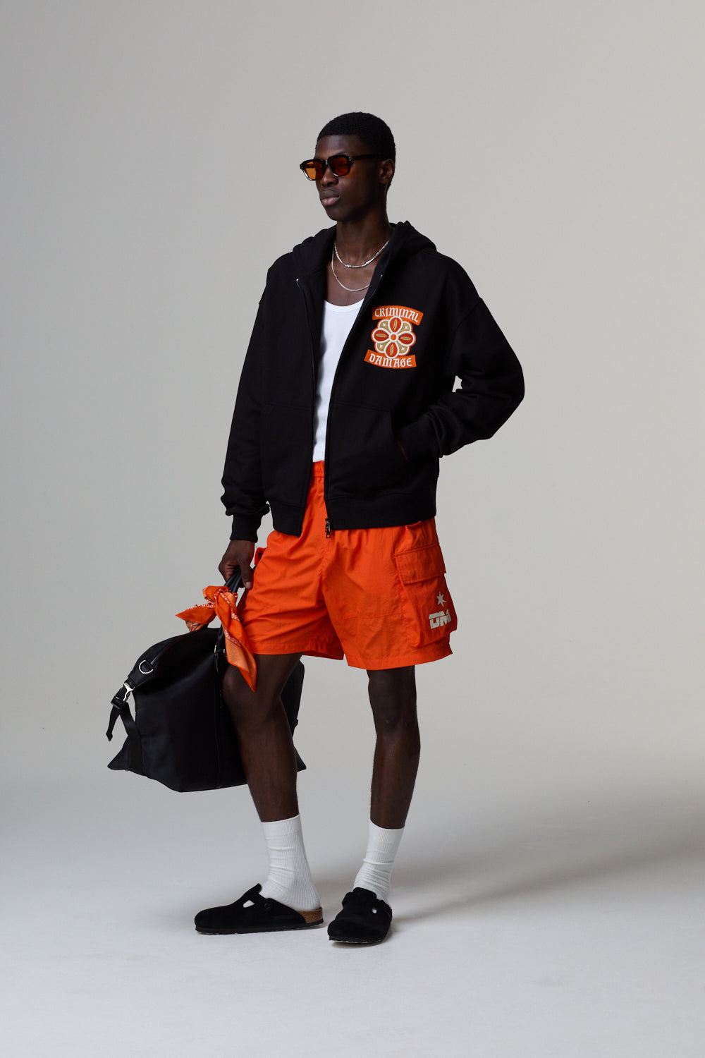 Model wearing a black jacket with an orange logo and orange shorts, holding a black bag on a light grey background.
