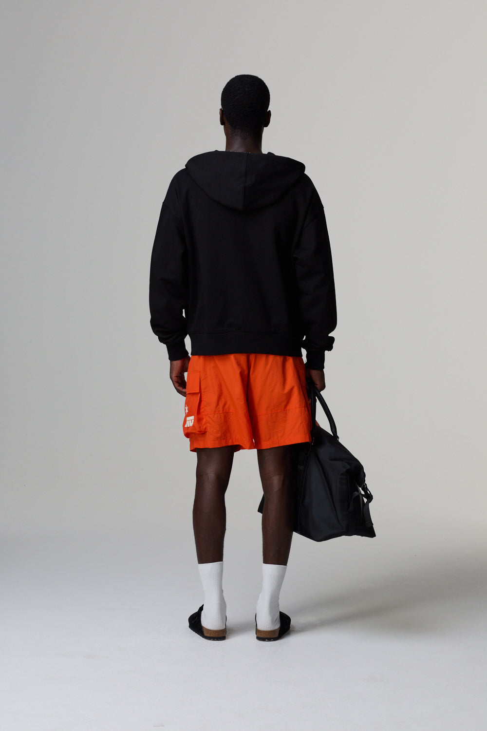 Model wearing a black Criminal Damage hoodie and orange shorts, holding a black bag on a grey background