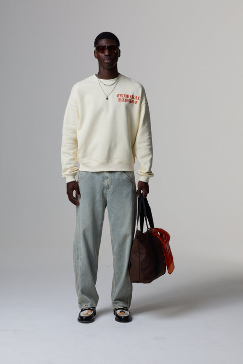 Model wearing a cream Criminal Damage sweatshirt with text, light grey pants, and holding a brown bag on a grey background