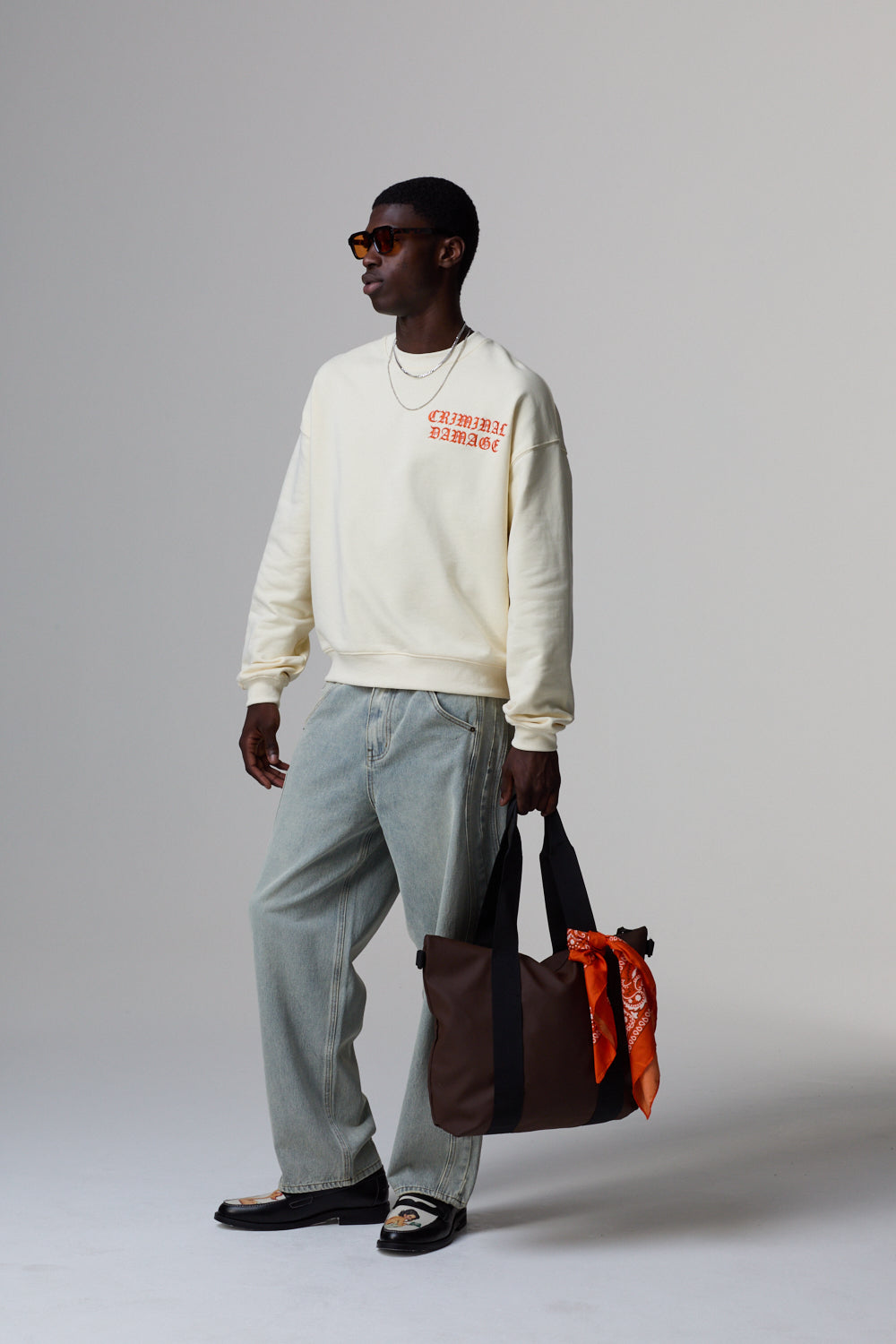 Model wearing a cream Criminal Damage sweatshirt with text, light blue jeans, and holding a duffle bag against a grey background