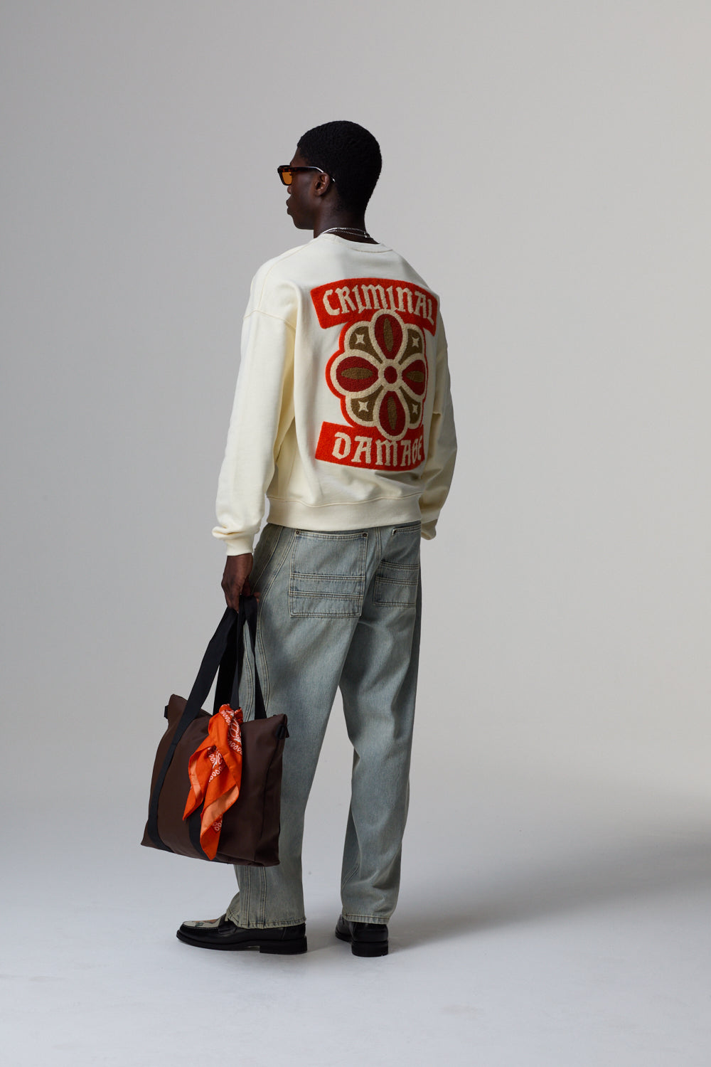 Model wearing a cream Criminal Damage sweatshirt with a red graphic design on a grey background