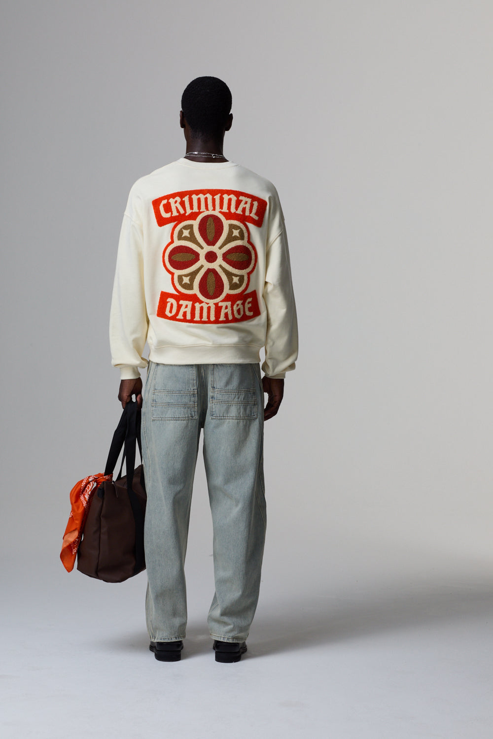 Model wearing a cream jacket with 'Criminal Damage' design, holding a brown bag on a grey background