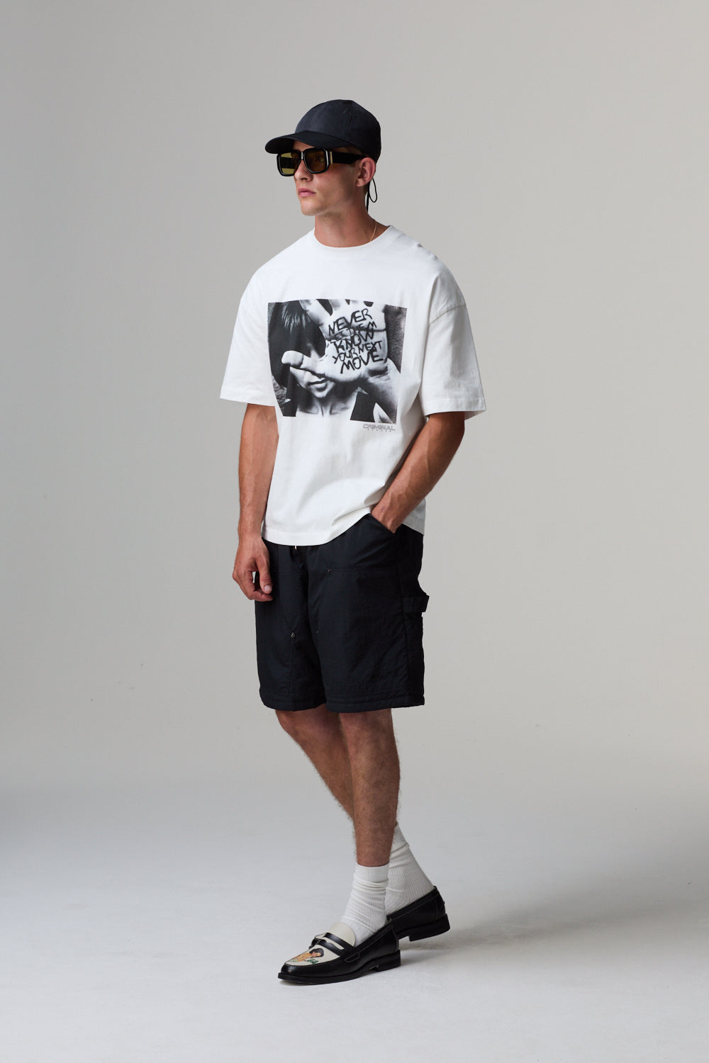 Model wearing a white Criminal Damage t-shirt with a graphic design, black shorts, and a cap on a grey background