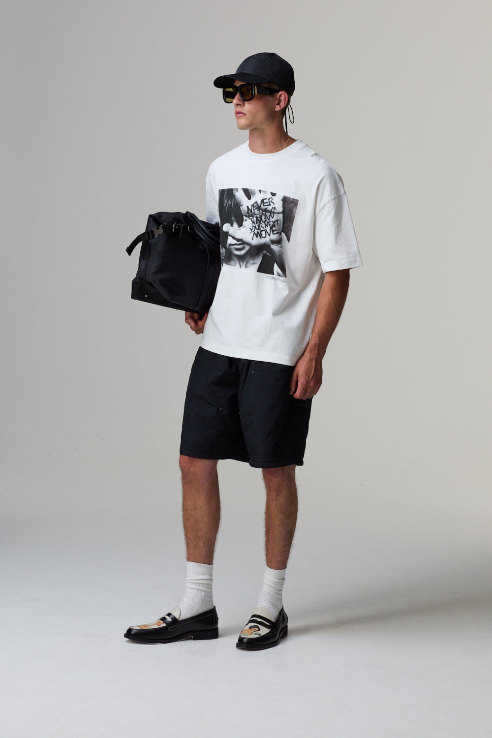 Model wearing a white Criminal Damage t-shirt with a graphic design, black shorts, and a cap, holding a black bag on a grey background.